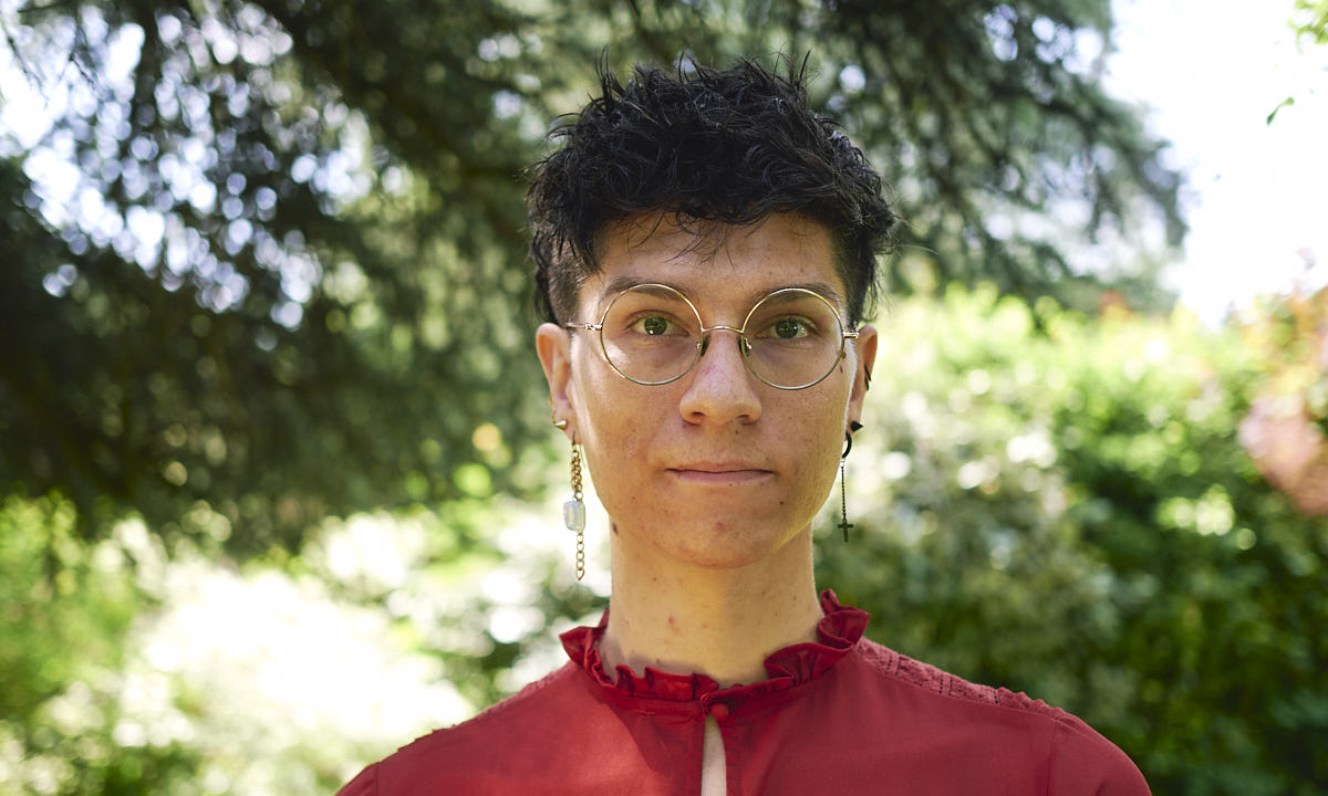 An androgynous white person with short black hair, round glasses and ear jewellery.