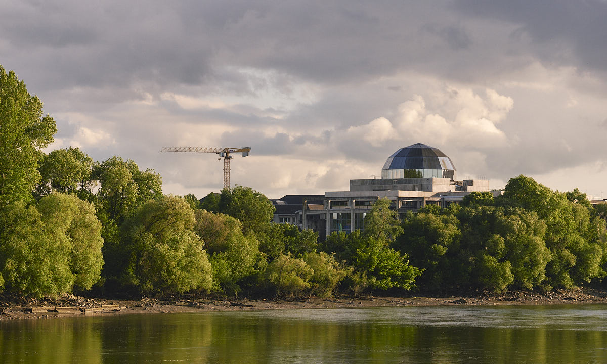 Building with a reflective dome hidden behind trees, with a river next to it and dramatic clouds
