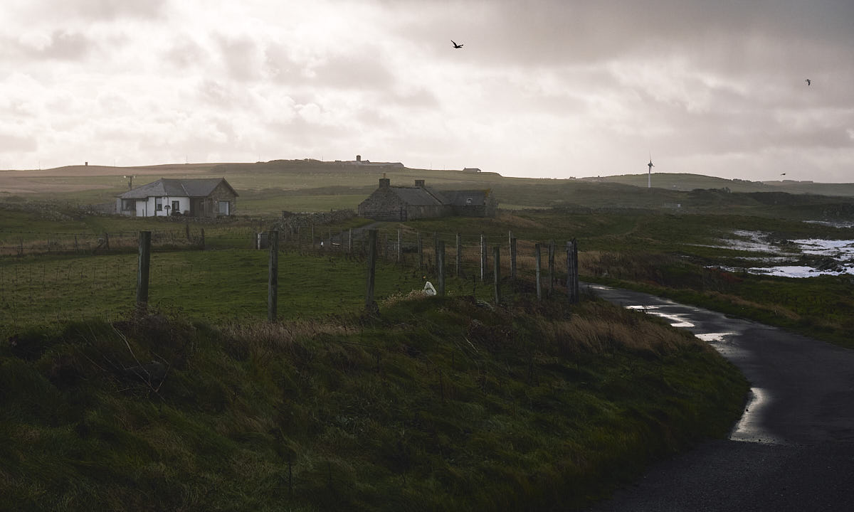 Road leading into grassy hills, with moody skies and foggy light. A few rural houses and a wind turbine can be seen